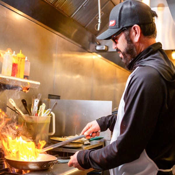 Chef in a black cap and apron flambéing food in a pan with large flames.