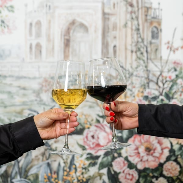 Two hands clinking wine glasses, one red and one white, against a floral and Indian-inspired mural.