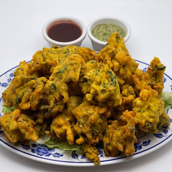 A plate of golden-brown Indian pakoras with two dipping sauces on a white background.