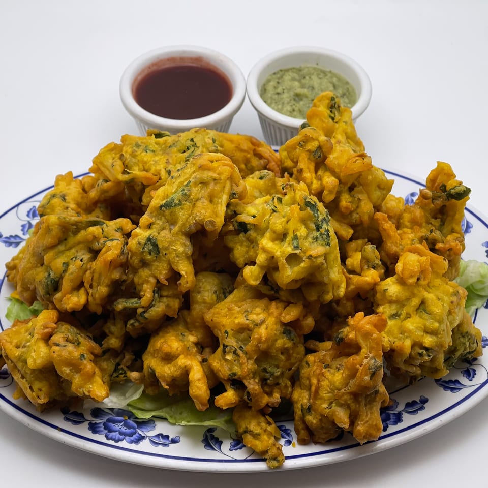 A plate of golden-brown Indian pakoras with two dipping sauces on a white background.